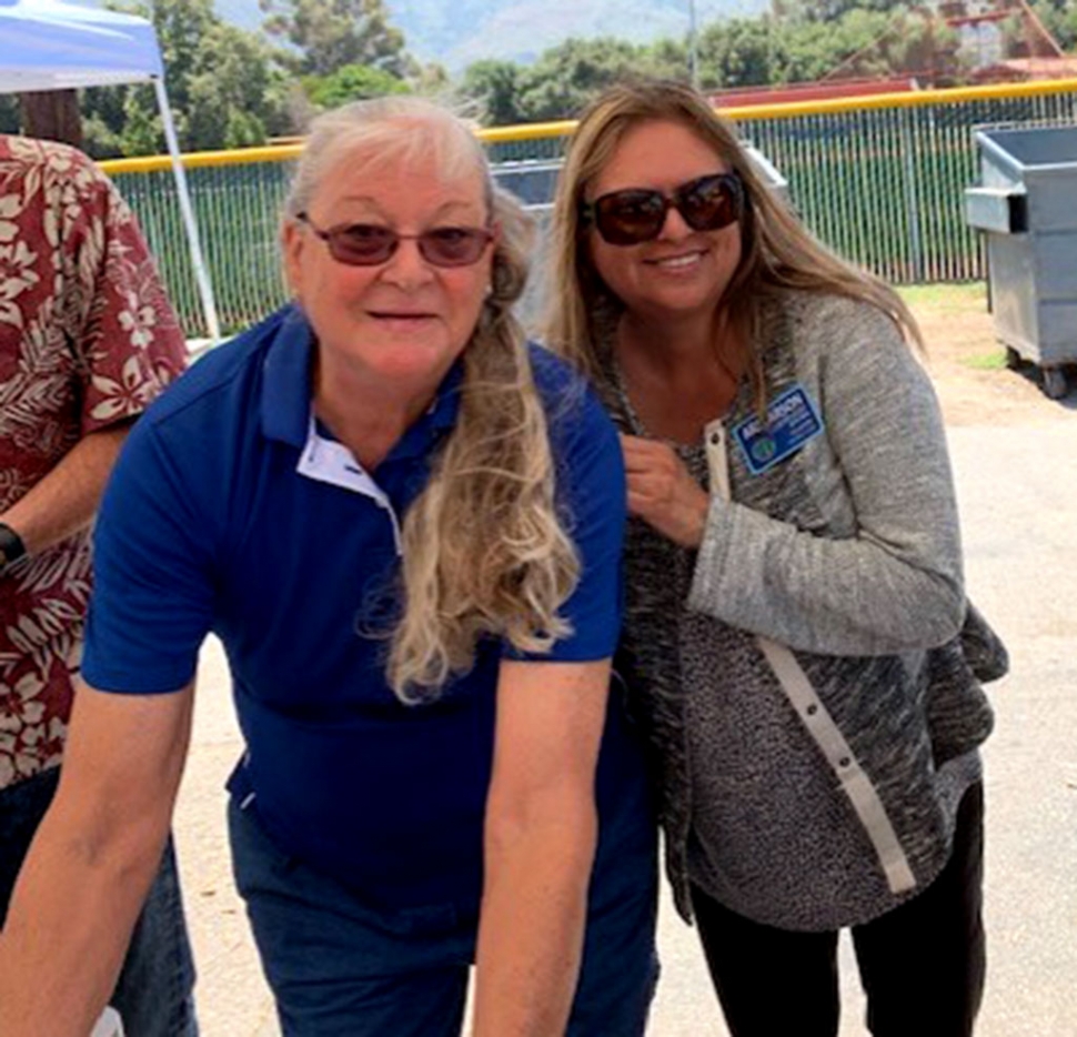 Rotary Member Cindy Blatt and President Ari Larson smiling for a photo as they help hand food and books to the kids.